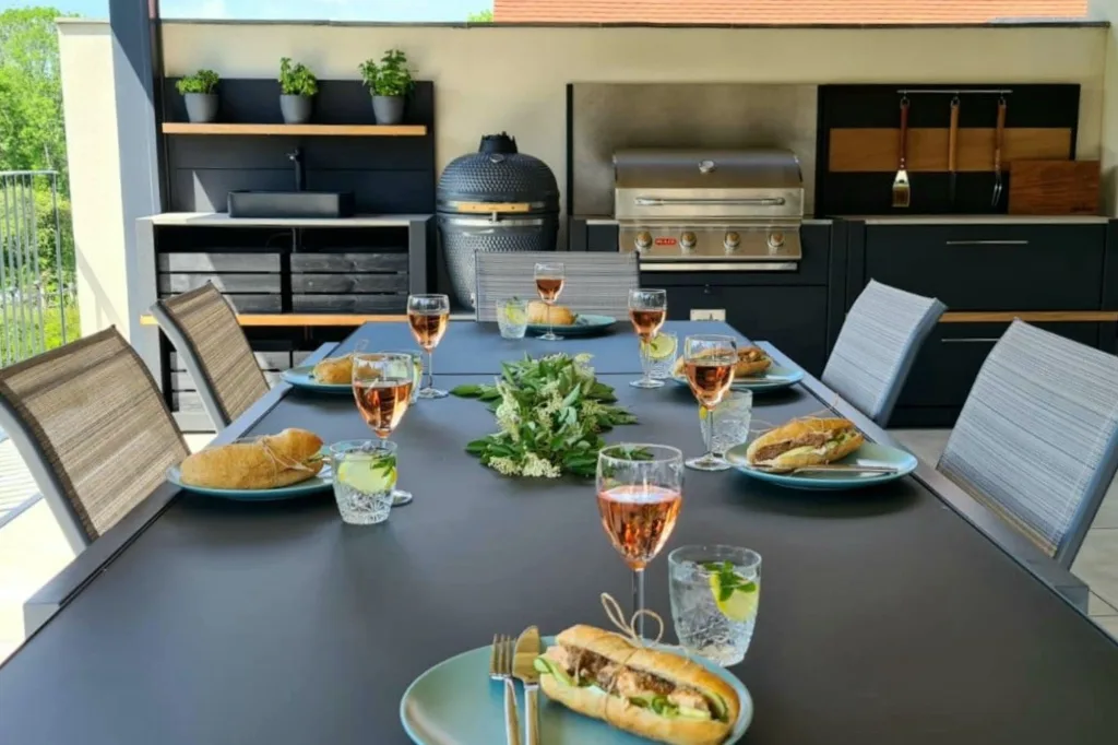 A lovely outdoor dining spread in front of the outdoor kitchen in this project. Featuring glasses of wine, baguettes on the table, along with a background of a Kamado BBQ, a gas BBQ and outdoor kitchen worktops.