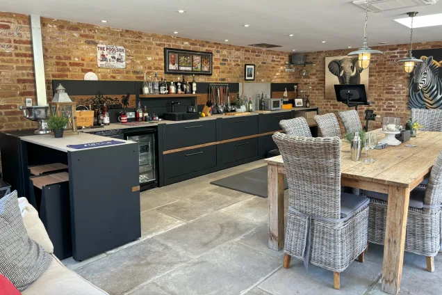Outdoor kitchen in saffron walden with storage units and a drinks fridge and dining table with chairs in garden building