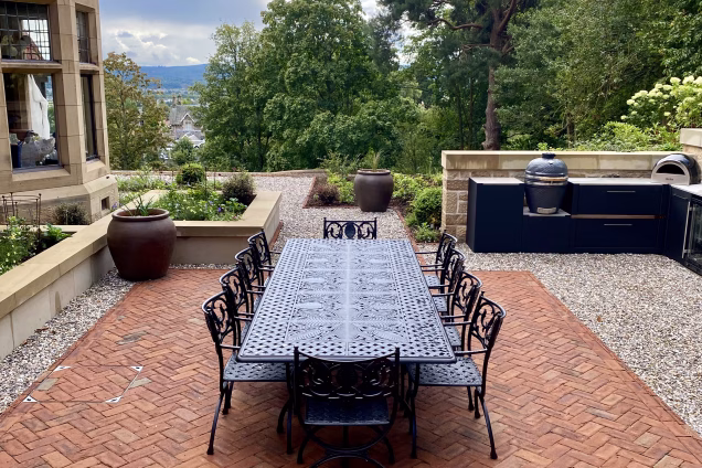 large outdoor table and chairs on large red paving section surrounded by stones with outdoor kitchen with gusto charcoal bbq