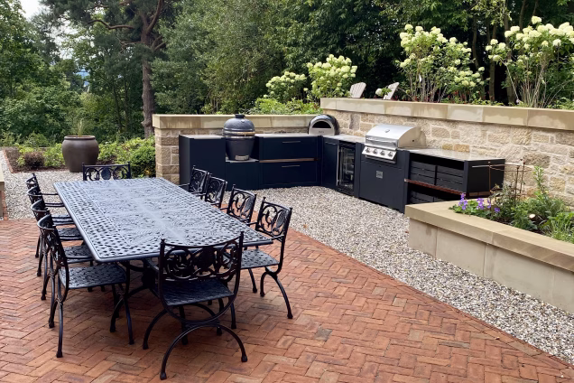 outdoor kitchen in garden with large trees, outdoor table, planters and three bbq cooking options with storage units