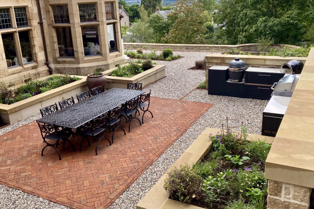 outdoor kitchen in large outdoor living area with table and chairs on red paving and planters
