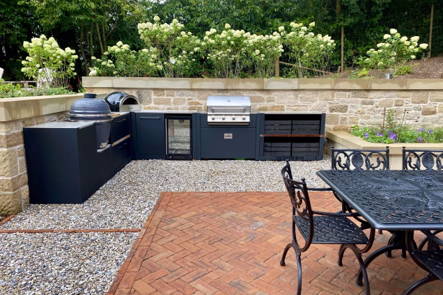 outdoor kitchen on stones next to planter with outdoor table and chairs on red paving blocks
