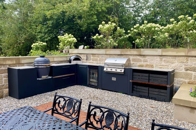 outdoor kitchen in Scotland in front of stone wall next to large outdoor table and chairs with three bbqs and storage and fridge and bin unit