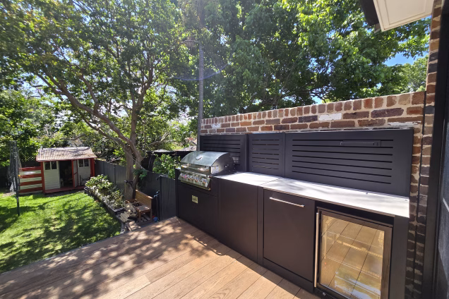 A view along an outdoor kitchen into a grassy, sunny backyard with a shed in Naremburn NSW. The outdoor kitchen is against a rustic brick wall and has a Bull gas grill and an outdoor fridge.