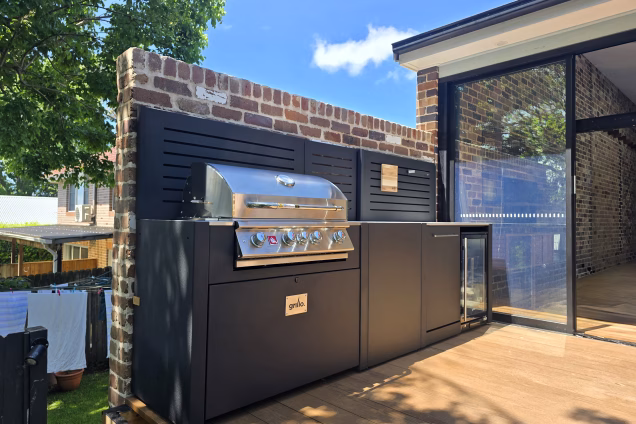 A Grillo outdoor kitchen on a raised deck against a rustic brick wall, with large glass patio doors at one end leading seamlessly to an open-plan living space with a matching brick wall