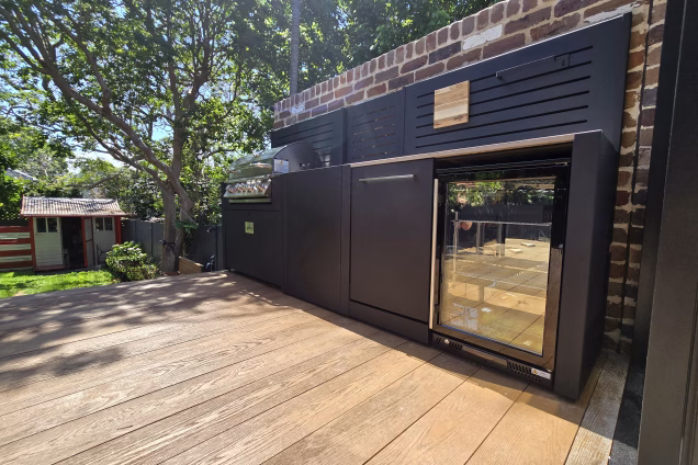 A low level wide angle photo focused on an outdoor kitchen on a raised deck, overlooking a Sydney garden on a sunny day