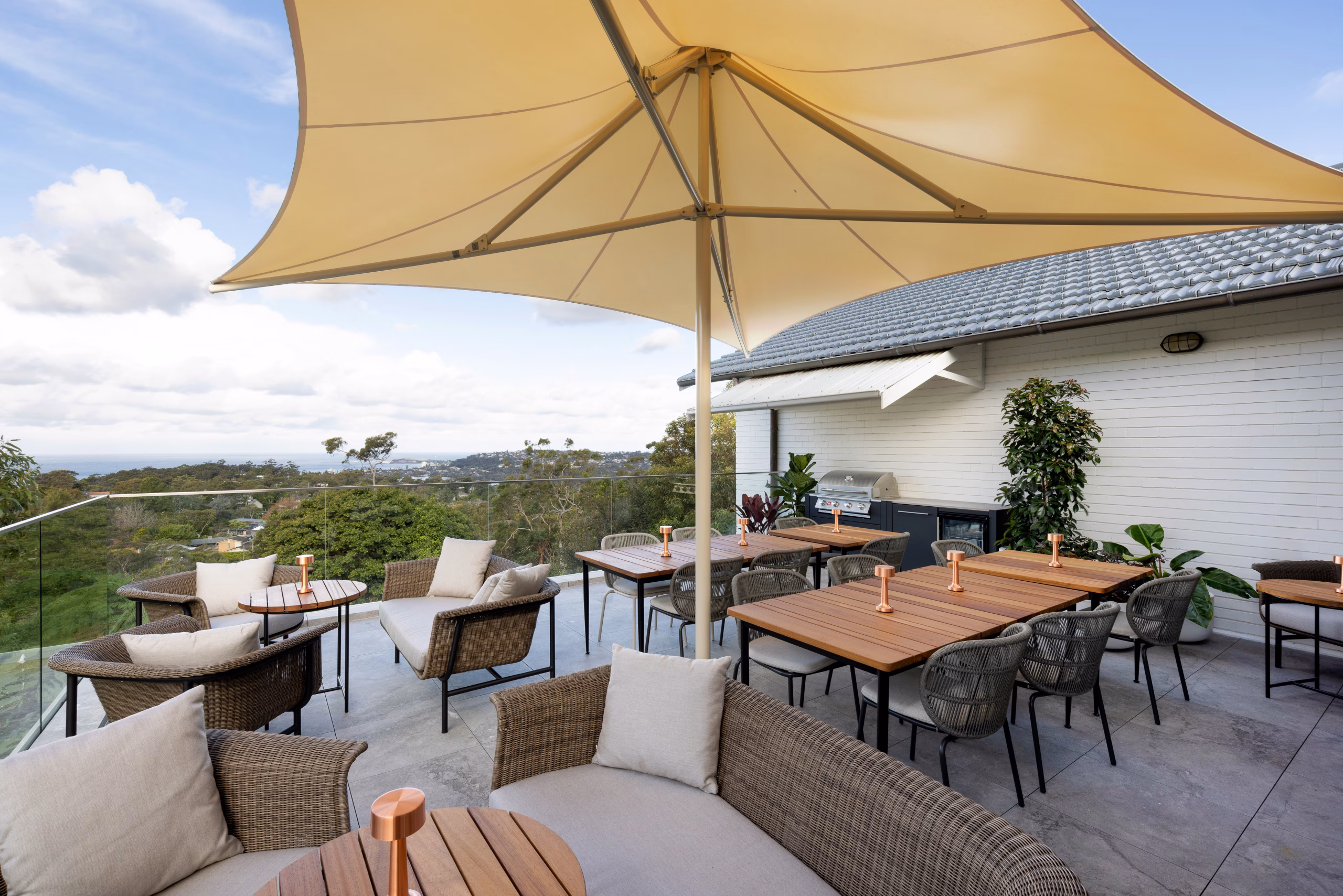A wide angle view under a sunshade on The Verandah, with seating and a Grillo kitchen in the background, showing the Bull gas grill and outdoor fridge