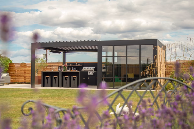 A modern outdoor kitchen and lounge area sits in a spacious garden, featuring a sleek black pergola with bar seating and a built-in grill. Behind it is a contemporary garden room with large glass panels.