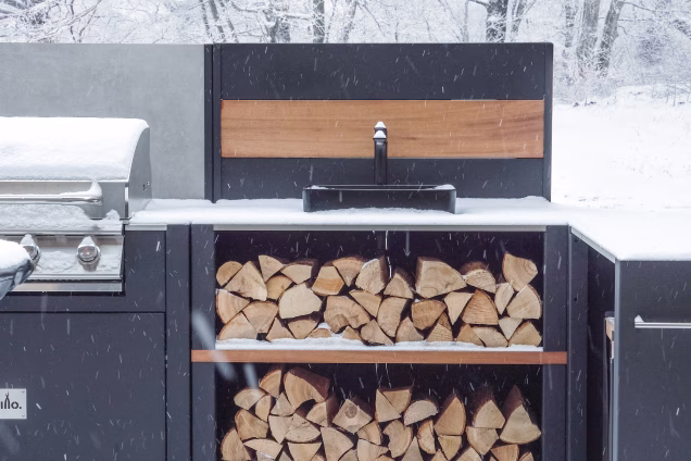 Grillo Vantage outdoor kitchen in heavy snow, with a gas grill to one side and a sink module centre of photograph, and snowy logs on the shelf underneath