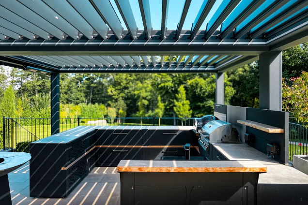 This contemporary outdoor kitchen boasts a state-of-the-art gas BBQ and a pizza oven, perfect for culinary enthusiasts. A sleek bar area invites guests to gather and enjoy refreshments, while a generous outdoor living space beneath a stunning, luxurious pergola creates an inviting atmosphere for relaxation and entertainment.